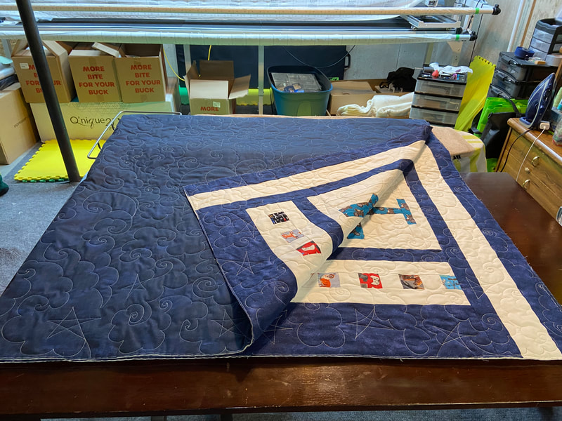 A Queen size quilt is folded into quarters. The dark blue backing is mostly visible with stars, clouds, swirls, and moons stitched on the fabric. One corner is turned up revealing part of the front. There is a partial blue and white border with small squares of colourful print fabric and an airplane.
