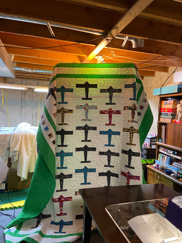 Queen size quilt with airplanes in red, blue, yellow, and black fabrics. The rows of airplanes are borders by green and white fabric. The left and right side borders have small squares of matching fabric to the airplanes creating an airport runway effect. 