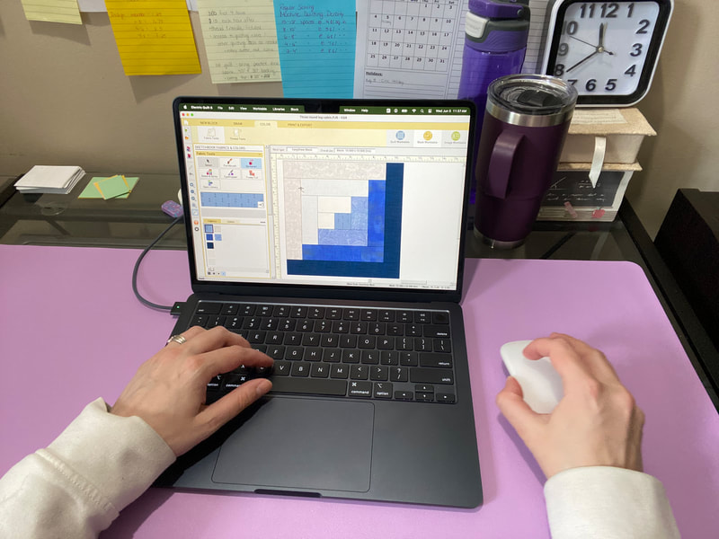 A lap top screen is displaying a log cabin quilt block in the EQ8 software. There are two hands in the photo working on the project. The laptop is positioned on a desk in a home office. 