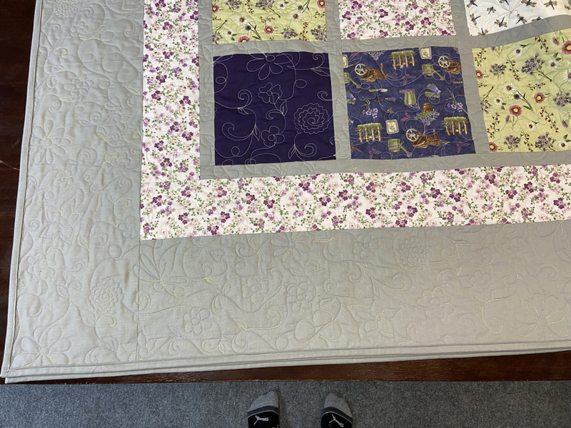A flower garden motif containing roses, poppies, daisies, plant tendrils with leaves, dragon flies, and butterflies is quilted on a grey, pink, green, and purple quilt. There is a grey border and pink flower print border. The body of the quilt is large print squares with flower and garden themes framed with grey sashing.